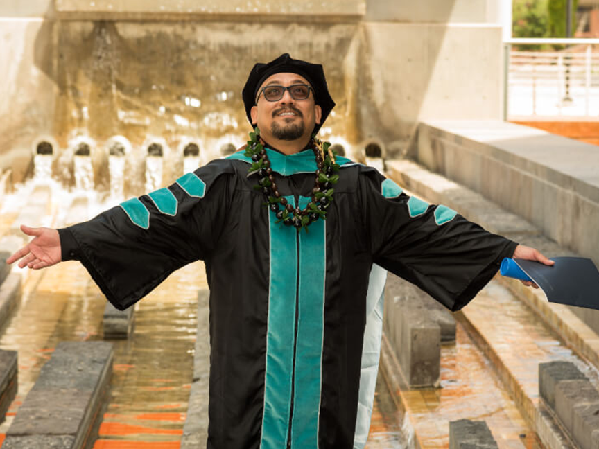 ATSU DPT graduate in his gown, holding his diploma in one hand with arms outstretched, looking up at the sky