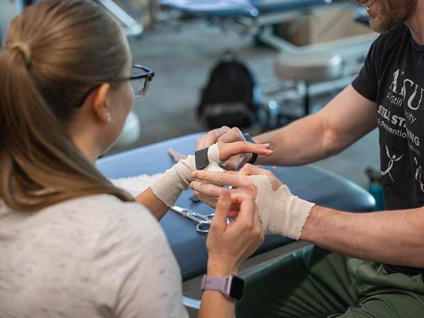 MSOT student practicing bandage wrapping on a classmate's hand, demonstrating hands-on training in occupational therapy skills and techniques.