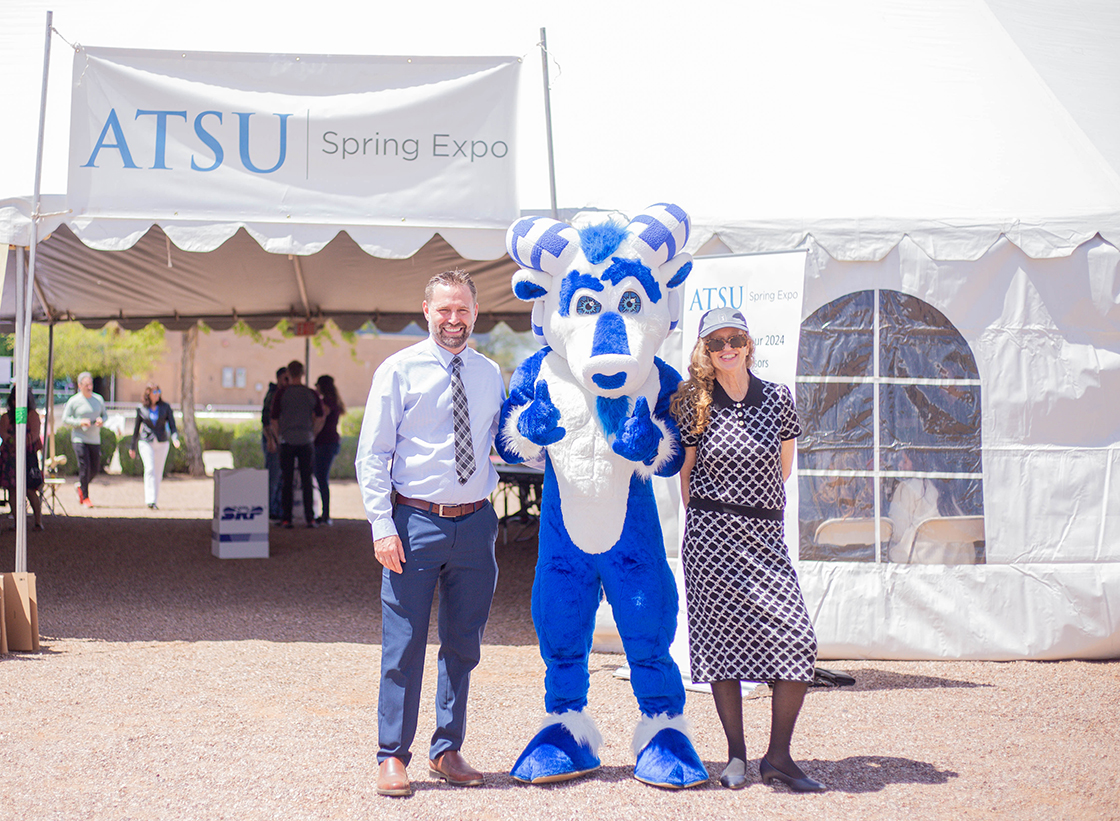 Two people stand with a mascot outside of a tented area